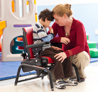 A therapist places her head on the head of a boy with disabilities who is sitting with support in an Activity chair helping him use problem solving skills for cognitive development 