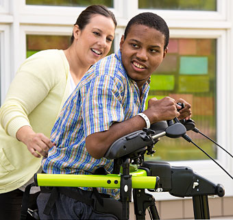 A young boy looks over his shoulder as a therapist guides him over-ground in a gait trainer