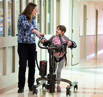 A therapist helps a student walk down the hall in a TRAM to the restroom for toilet transfers.