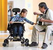 A man plays a guitar and sings to help a boy in a special needs chair focus on the task he is performing