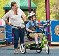 A young boy in a blue shirt and shorts rides on a green adaptive bike as his therapist helps to push him for functional mobility exercise.