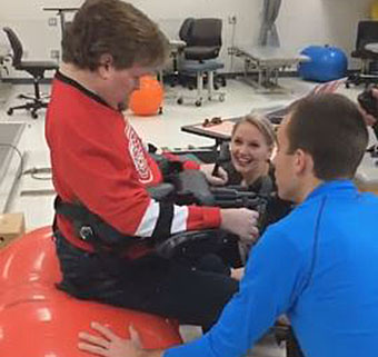 A man with disabilities sits on a red therapy ball while PT students demonstrate a creative mobility technique.