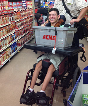 A young man in special education rides in a wheelchair and holds a gray shopping basket as he looks for ingredients for a cooking project in his life skills class.