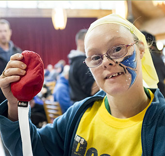 A young woman wearing a yellow Rifton T-shirt smiles as she holds up a red bean bag at rifton's birthday party.