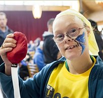A young woman wearing a yellow Rifton T-shirt smiles as she holds up a red bean bag at rifton's birthday party.