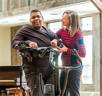 A therapist smiles at a young man standing in the Rifton TRAM patient lift acquired with a workers compensation grant.