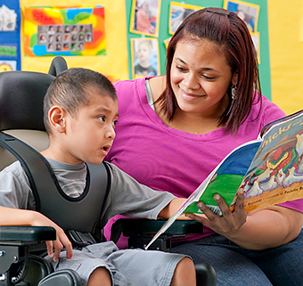 A caregiver reading a picture book to a young boy sitting in a Rifton Activity Chair.