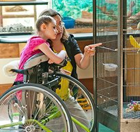 A young girl standing in a Rifton Mobile Stander looks at a goldfinch in a cage with her caregiver.