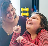 A caregiver and teenage client laughing together in a gym during a therapy session.