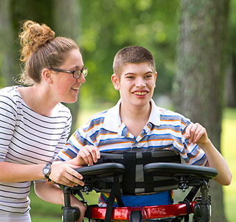 A teenage boy walks outside in a Rifton Pacer Gait Trainer while an aid assists him.
