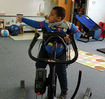 A young boy uses the rifton tram for safe transfers to standing and ambulation over the carpet of the gym.