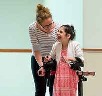A girl in a pink dress smiles at her caregiver, while walking in her Rifton Pacer gait trainer.