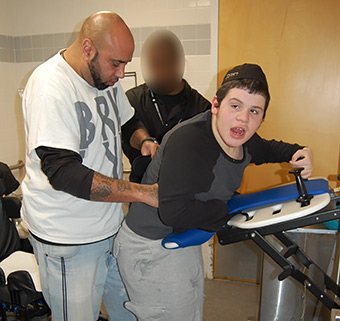 A young boy stands at a Rifton Support Station with the aid of a caregiver.