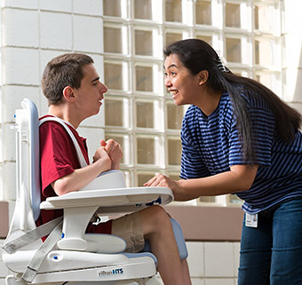 A young female talks to a young boy sitting in a Rifton HTS.