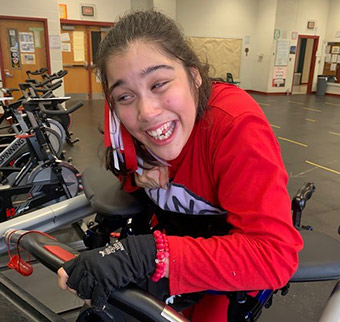 A teenage girl with cerebral palsy stands on a treadmill in a gym.