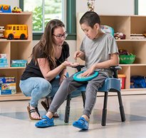 A caregiver talks to a boy sitting in a Rifton Compass Chair in a classroom.