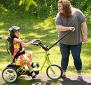 A girl sitting on a Rifton Adaptive Tricycle with a caretaker holding the front guide bar