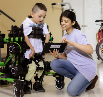 A little boy using a Trexo with a Rifton Pacer, while a PT shows him a tablet