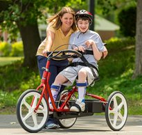 A boy riding a Rifton Adaptive Tricycle, with his caretaker standing next to him