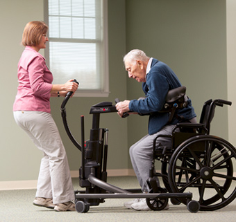 An elderly man stepping out of a wheelchair into a Rifton TRAM with the assistance of a caretaker