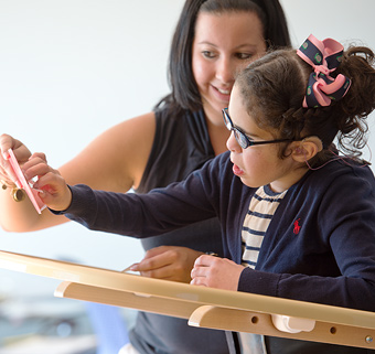 A girl standing in a Rifton Supine Stander, looking at a card with her caretaker