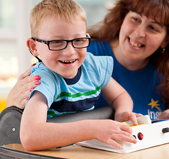 A boy standing in a Rifton Prone Stander, using a communication device with his therapist.