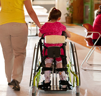 A young girl wheeling down a hallway in a green Rifton Mobile Stander, with a caretaker next to her
