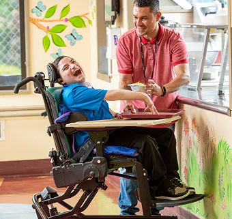 A boy sitting on a Rifton Activity Chair, with his therapist standing next to him, smiling at each other