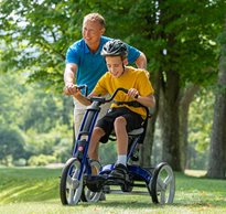 A boy pedals a blue Rifton Adaptive Tricycle, while his therapist walking beside him.