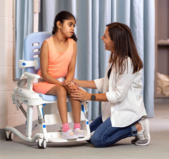 A young girl sits on a Rifton HTS (Hygiene & Toileting System) while her therapist helps her position.