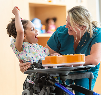 A girl stands in a Rifton Stander and smiles at her therapist.