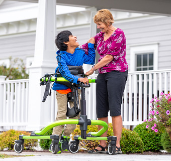 A boy walks in a Rifton Pacer Gait Trainer and smiles at his caretaker.