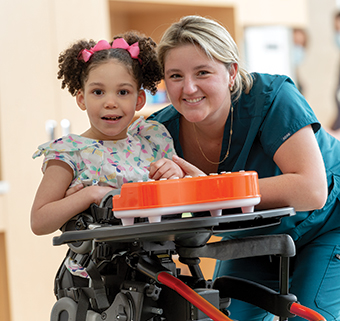 A girl stands in a Rifton Stander in prone configuration and smiles with her therapist.