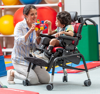 A boy sits in a Rifton Activity Chair and plays with a toy train, while his caretaker smiles.