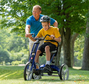 A boy pedals a blue Rifton Adaptive Tricycle, while his therapist walking beside him.