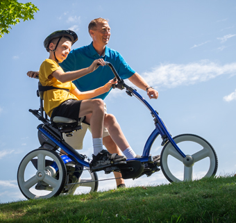 A boy rides on a Rifton Adaptive Tricycle next to his caregiver.