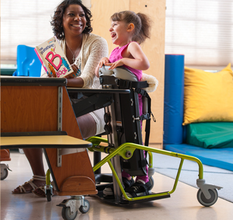 A girl stands in a Rifton Mobile Stander and looks at a book with her caretaker.