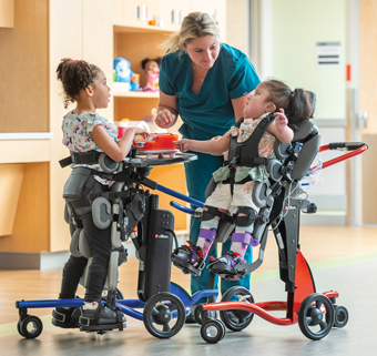 2 girls stand in Rifton Standers and play with their caretaker.