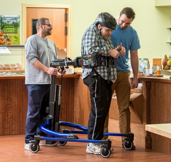 A man walks in a Rifton E-Pacer, with the assistance of two caretakers.