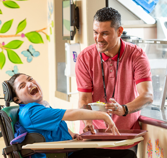 A boy sits in a Rifton Activity Chair while his caretaker serves him lunch.