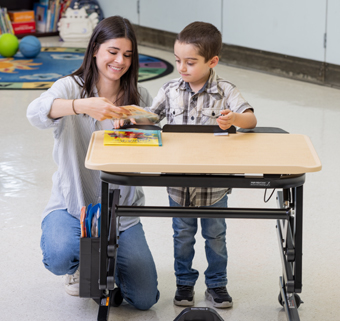 A boy looks at a book on a Rifton Adaptive Desk with his therapist.