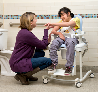 A girl sits on a Rifton HTS and smiles at her caretaker.