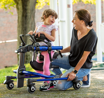 A girl stands in a Rifton Pacer and smiles at her caretaker.