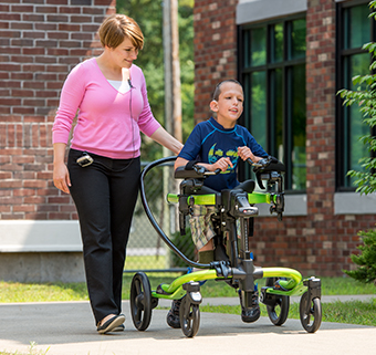 A boy walks in a Rifton Pacer on an outdoor path, accompanied by a therapist.