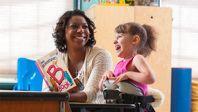 A girl stands in a Rifton Mobile Stander and reads a book with her caretaker.