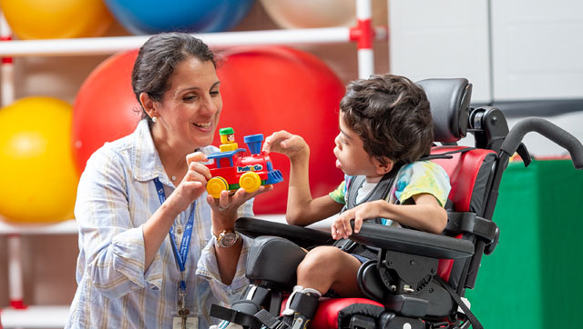 A boy sits in a Rifton Activity Chair and plays with a toy train with his caretaker.
