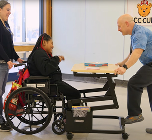 A young woman sits in a wheel chair while a therapist adjusts a Rifton Adaptive Desk for her.