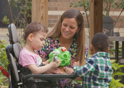 A girl stands in a Rifton Stander and plays with a boy and their caretaker.
