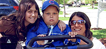 Scott Conlon, a school-aged boy, with his mother an older sister, out on the street riding his Rifton adaptive trike.