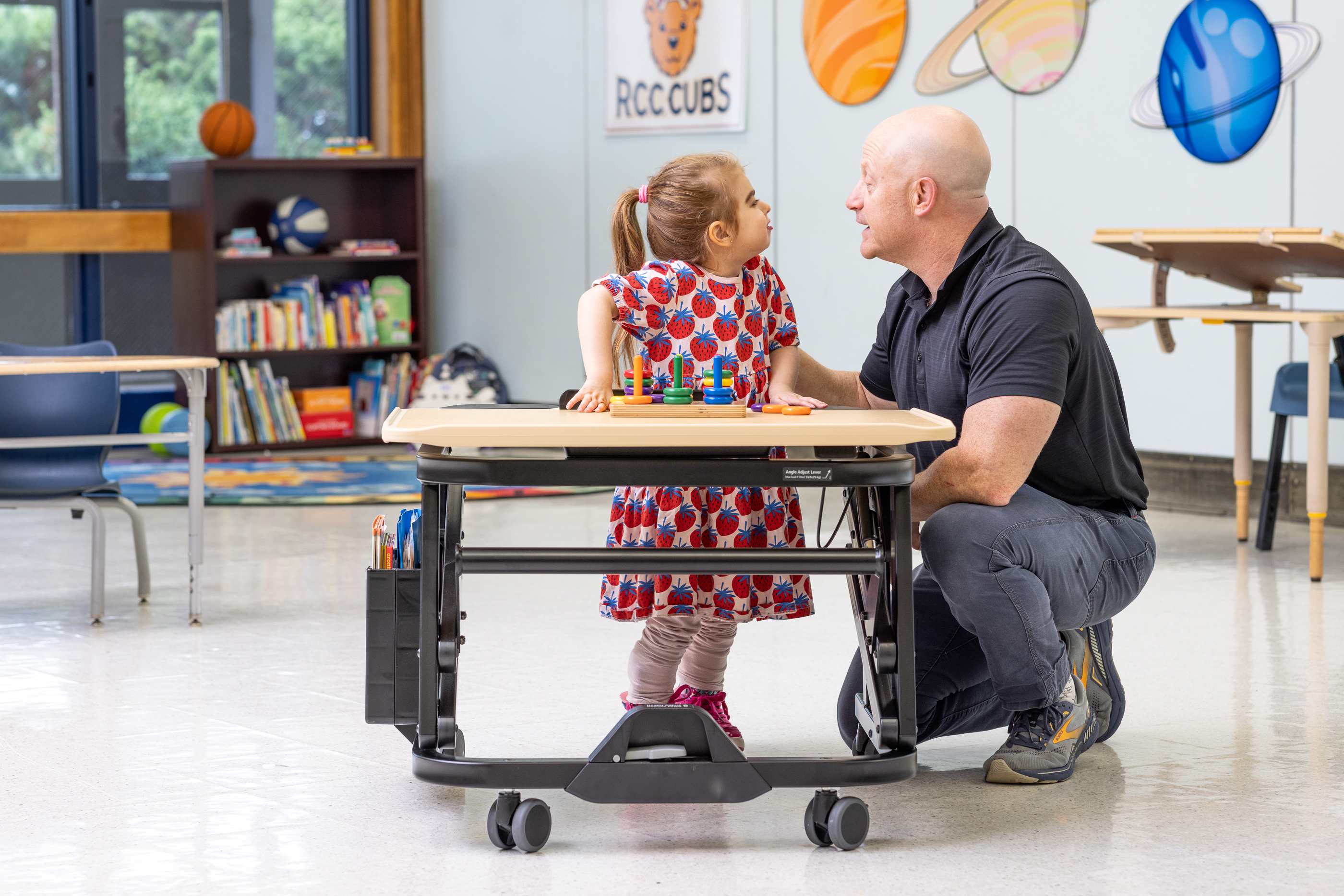 A girl stands by a Rifton Adaptive Desk and smiles at her therapist.
